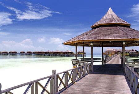 Image of a walkway and huts on stilts on a remote Malaysian tropical island with deep blue skies and crystal clear waters.の写真素材