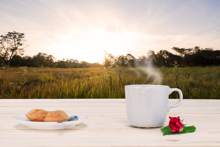 Hot coffee with steam, dessert and red flower with green leaf  on vintage wooden table top on blurred meadow during sunrise in morning with flare backgroundの写真素材