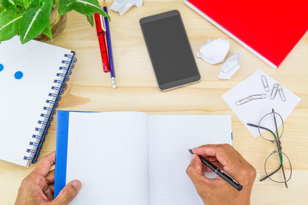 Hand writing on notebook on wooden desk with crumpled paper, office supplies, smartphone, glasses...