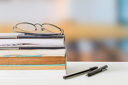 Eyeglasses on books and pens on white table on blurred library backgroundの写真素材