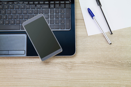 Smartphone on laptop with white paper and pens on wooden desk, business conceptの写真素材