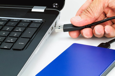 Hand connecting external hard drive usb cable to laptop on white desk