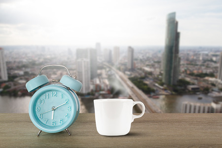 Blue classic alarm clock and hot white coffee cup with steam on vintage wooden desk on blurred city backgroundの写真素材