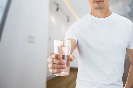 Man wearing white shirt holding glass of drinking water in a house, health care concept