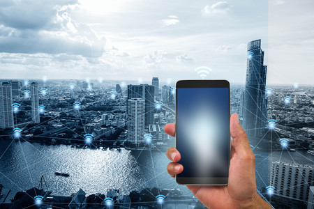 Hand holding mobile phone on blue tone smart city with wifi network connections background, communication network technology conceptの写真素材