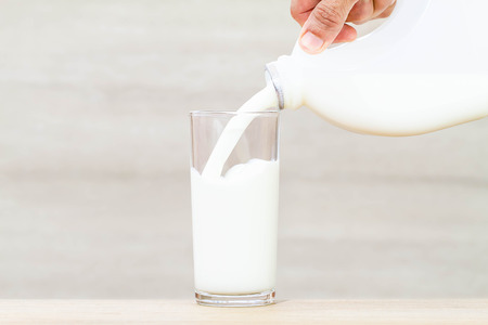 Hand holding bottle of fresh milk pouring into drinking glass on wooden tableの写真素材