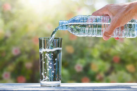 Hand holding drinking water bottle pouring into glass on blurred green nature with flowers backgroundの写真素材