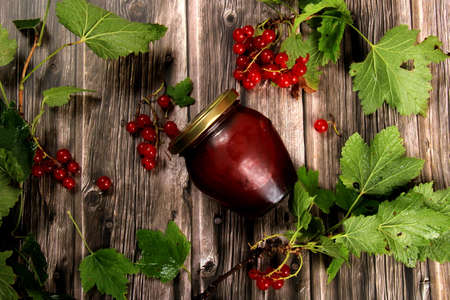 Glass jar with berry jam or sauce. red currant branches with green leaves on a background of wooden boardsの写真素材