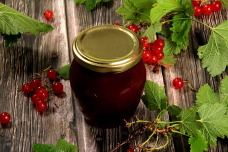 Glass jar with berry jam or sauce. red currant branches with green leaves on a background of wooden boardsの写真素材