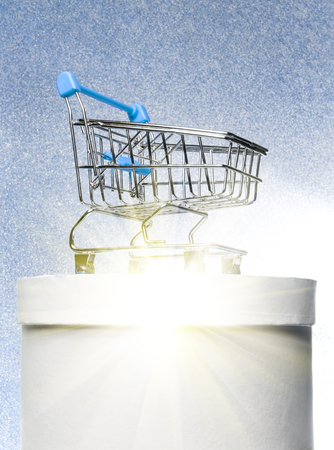 Shopping trolley on a round white gift box on a winter blue shiny background with bright rays.の写真素材