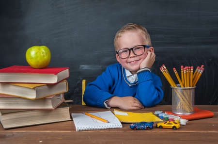 Funny little child sitting on a desk with stack of books and apple. back to school written on chalk blackboardの写真素材