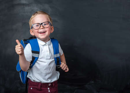 Happy smiling boy in glasses with thumb up is going to school for the first time. Child with school bag and book. Kid with blackboard on a background. Back to schoolの写真素材