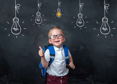Back to school. Funny little boy in glasses pointing up on blackboard. Child from elementary school with book and bag. Education. Bulb with light on blackboard. Thinking smart kidの写真素材