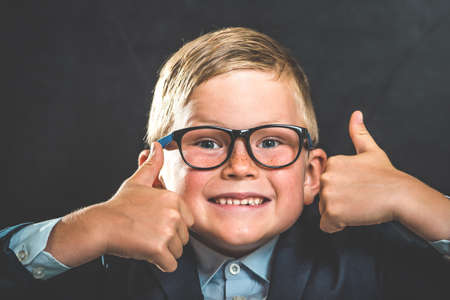 Portrait of smart school child with thumb up near blackboard blank and looking at camera. Happy kid boy in uniform. Go back to school. First day of fall. Funny faceの写真素材