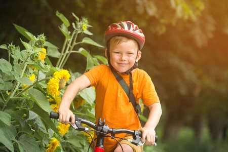 Portrait of a cute boy on bicycle. Happy smiking kid on bike. Child in safety helmet and orange shirt playing outdoors in park. Sport and healthcare concept. School kid in bike. Safetyの写真素材