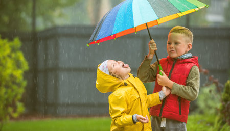 Little child girl and boy playing under autumn shower. Happy time for children in park. Kid catching rain drops. Bright style clothes rainbow umbrella and warm duffle coat. Health Care and protectionの写真素材