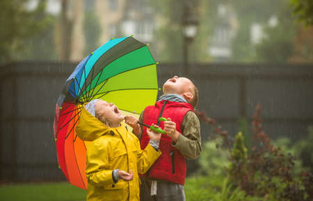 happy kids catching raindrops in park. Little boy and girl under colorful umbrella. Happy childhood concept. Bright style clothes in autumn weatherの写真素材