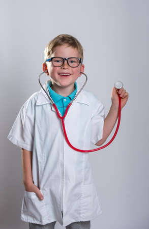 Boy playing doctor with a stethoscope. Kid in glasses isolated over white. Thumb up. White clothesの写真素材