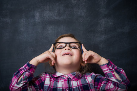 Cute girl is holding glasses and looking up, isolated over blackboard background. Innovation. Idea. Dream. Thinkの写真素材