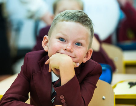 School kid in class. funny little boy pupil smiling while sitting in desk in suit. inquisitive,...
