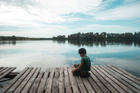 Boy sitting alone on pier while fishing from dock on lake. Kid on beautiful morning in natureの写真素材