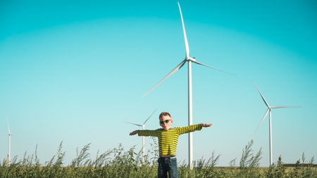 child playing with the wind near a wind turbine. Wind mills. Kid boy in sunglasses with hands like a plane. Renewable energies and sustainable resources - wind mills.の写真素材