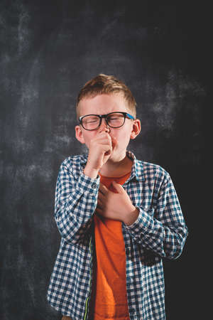Cute little boy is coughing. Little school kid in glasses against blackboard.の写真素材