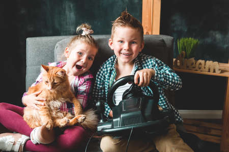 Funny little girl and boy playing game with steering wheel console. Funny kids with pet cat in hands sitting on sofa at home. Happy brother and sister. Family time.の写真素材