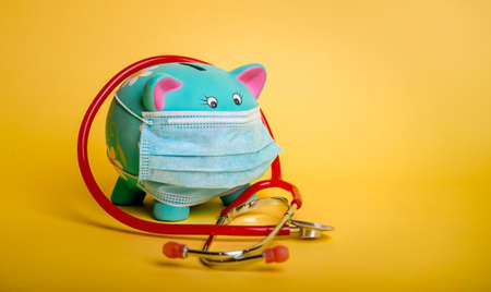 Stethoscope around blue Piggybank isolated on yellow background . Piggy bank in protective medical face mask.の写真素材