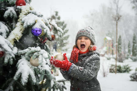 Portrait of happy girl looking in camera while decorating Christmas tree with toy balls. Winter weather. Christmas tree in snow outdoors. Stylish child in hat and coat and red scarf and mittensの写真素材