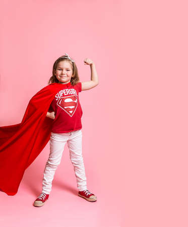 Super girl power. Kid in red t-shirt and cloak isolated on pink background. Confident child shows muscles. Kindergarten kid ready to go to schoolの写真素材