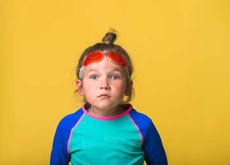 Portrait of funny little girl on yellow background. Kid in swimsuit and goggles for swimming. Emotional surprised kid face with big eyes looking in camera. Swim or dive in school or water parkの写真素材