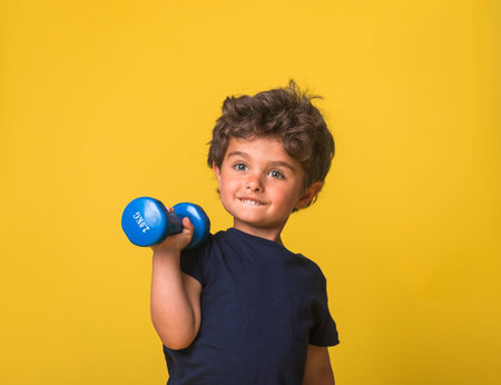 Portrait of funny little boy with dumbbells over yellow background. Healthy lifestyle for kindergarten and school kids. Sports and activities for children on summer vacationの写真素材
