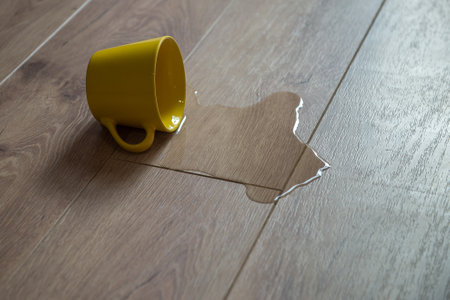 Yellow cup of water spilled on wooden laminate floor. Moisture protection of a floor. Parquet board with spilled water.の写真素材