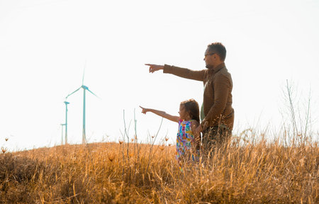 Mature father with small daughter standing on field on wind farm. Renewable energy and alternative electricity sources. Wind generators turbines. Concept of sustainable resources and peopleの写真素材