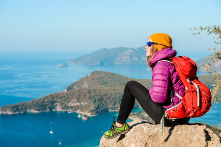 Young woman hiker in bright clothes using digital tablet sit at seaside cliff edge and enjoy the view at mountain peak. Beautiful islands sea and blue lagunes. Trekking route to travel aloneの写真素材