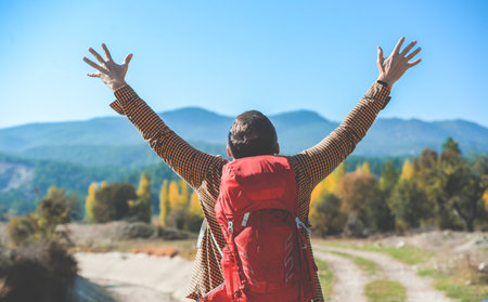 Rear view portrait of man with backpack raised hands looking on beautiful mountains view standing on a mountain. Hiking among beautiful nature views. Calm and excited with arms spread openの写真素材