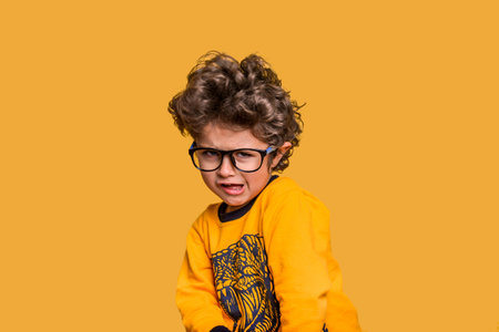 Portrait of five years old child making funny face. Boy in glasses tousled hair looking at camera with silly smile. Kid in yellow shirt isolated on yellow background. Hispanic cute childの写真素材