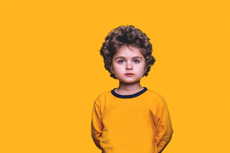 Funny little curly five years kid boy isolated on yellow background. Cute child looking in camera. Portrait of smart handsome serious kid. Childhood lifestyle concept. Kindergarten or school kidの写真素材