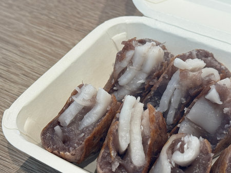 Close-up view of sliced traditional Thai coconut pancake filled with soft coconut meat and taro-like batter, served in an eco-friendly takeaway box on a wooden table.の写真素材