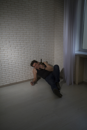 a drunken slovenly clad man lies and sleeps in the corner of the room and holds a bottle with a glass and tries to pour more alcoholの写真素材