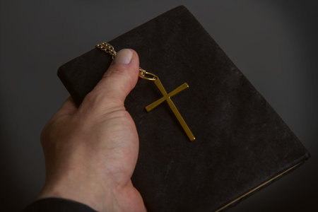 a man's hand holds a bible in a dark binder a metal cross on a chain close-up