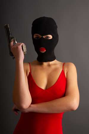 Young self-confident girl with a big gun in a black balaclava and in a red sexy dress standing on a gray backgroundの写真素材