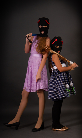 Two teenage hooligans in bright elegant dresses, but in gangster balaclava hats with heavy truncheons pose on a dark backgroundの写真素材