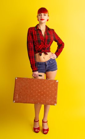 Pretty girl in knotted plaid shirt and shorts with a retro suitcase on a bright yellow backgroundの写真素材