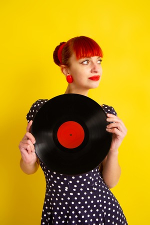 Pretty girl in retro dress in polka dot holding vinyl disc on a bright yellow backgroundの写真素材
