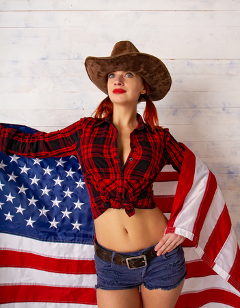 upright patriotic girl in a cowboy costume wearing a traditional wide-brimmed hat and shertach posing against a wooden background with the flag of the USA.の写真素材