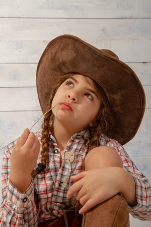 A little girl in a wide-brimmed cowboy hat wearing a traditional dress and high boots is eating a straw on a light wooden backgroundの写真素材