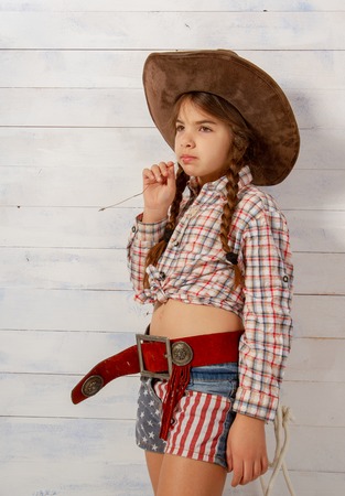 A little girl in a wide-brimmed cowboy hat wearing a traditional dress and with a lasso is eating a straw on a light wooden background.の写真素材