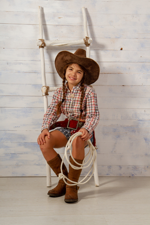 Little girl in a wide-brimmed cowboy hat and traditional dress in high boots and with a lasso posing on a light wooden backgroundの写真素材